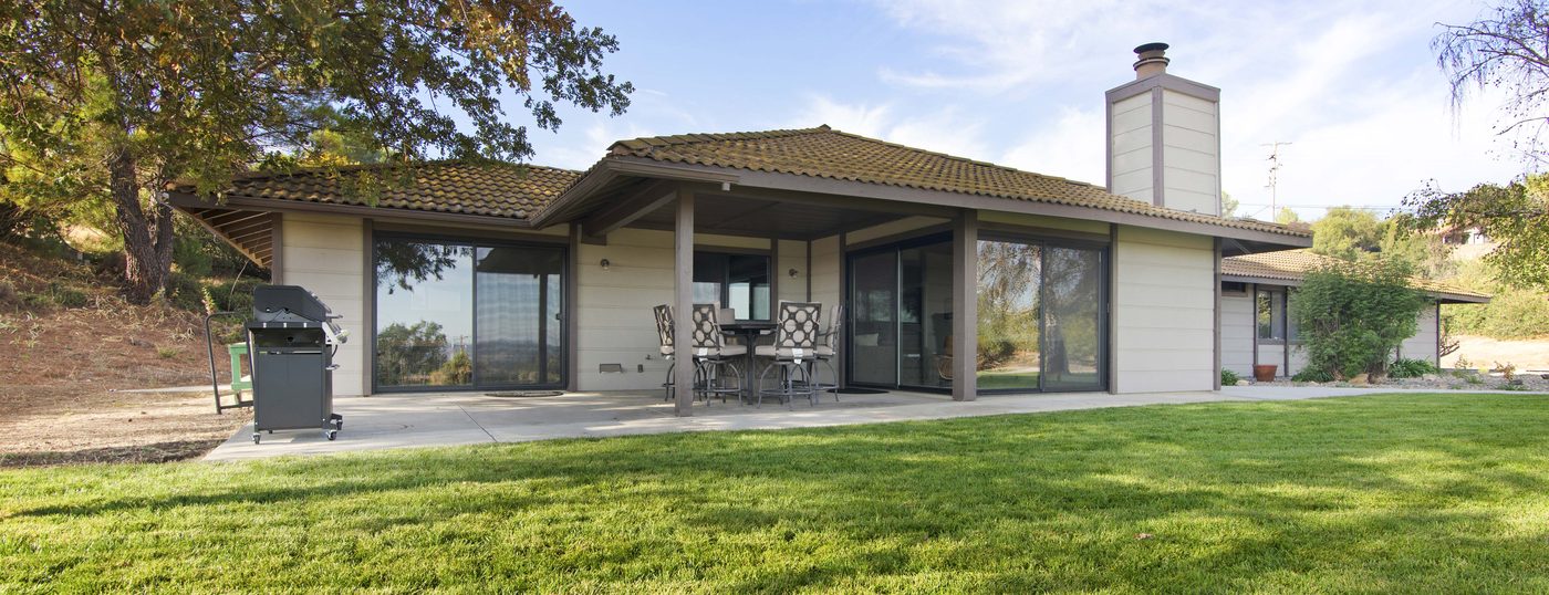 Single-story ranch-style home with a tile roof and deep covered patio, seen across a lawn beneath a mature oak